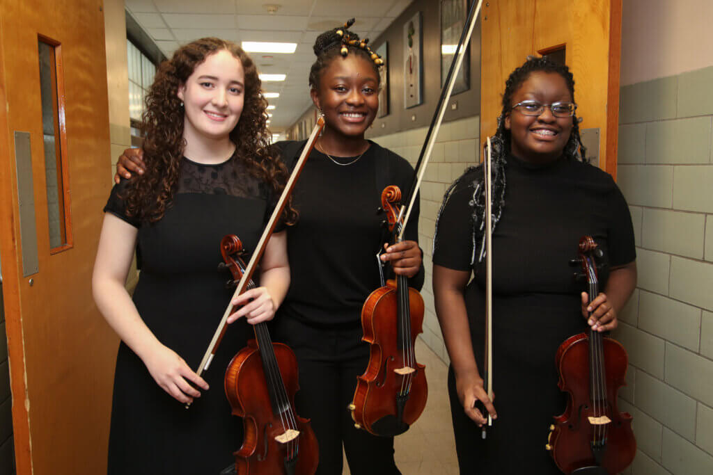 Three female students hold violins and smile for a photo.